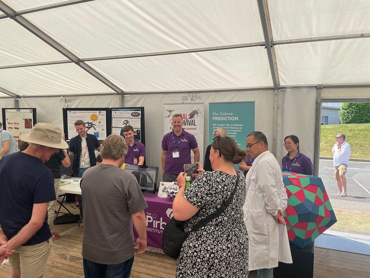 Group of Pirbright researchers in a marquee behind a table and in front of pull up posters with a small group of general public members looking at taking photos