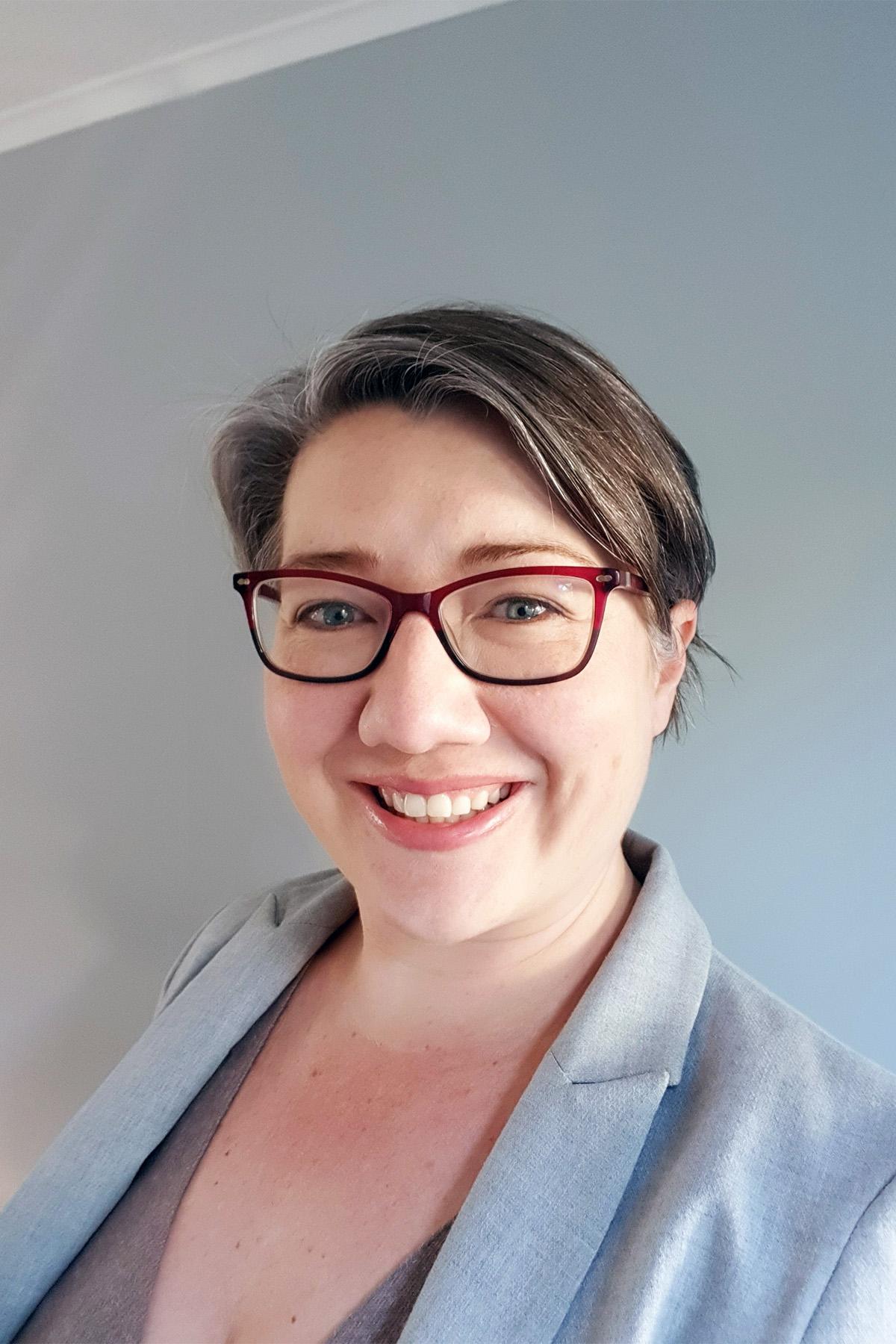 Lizelle Gouverneur smiles to camera - head and shoulders picture. She wears a light grey jacket, has red rimmed square glasses and short dark hair.