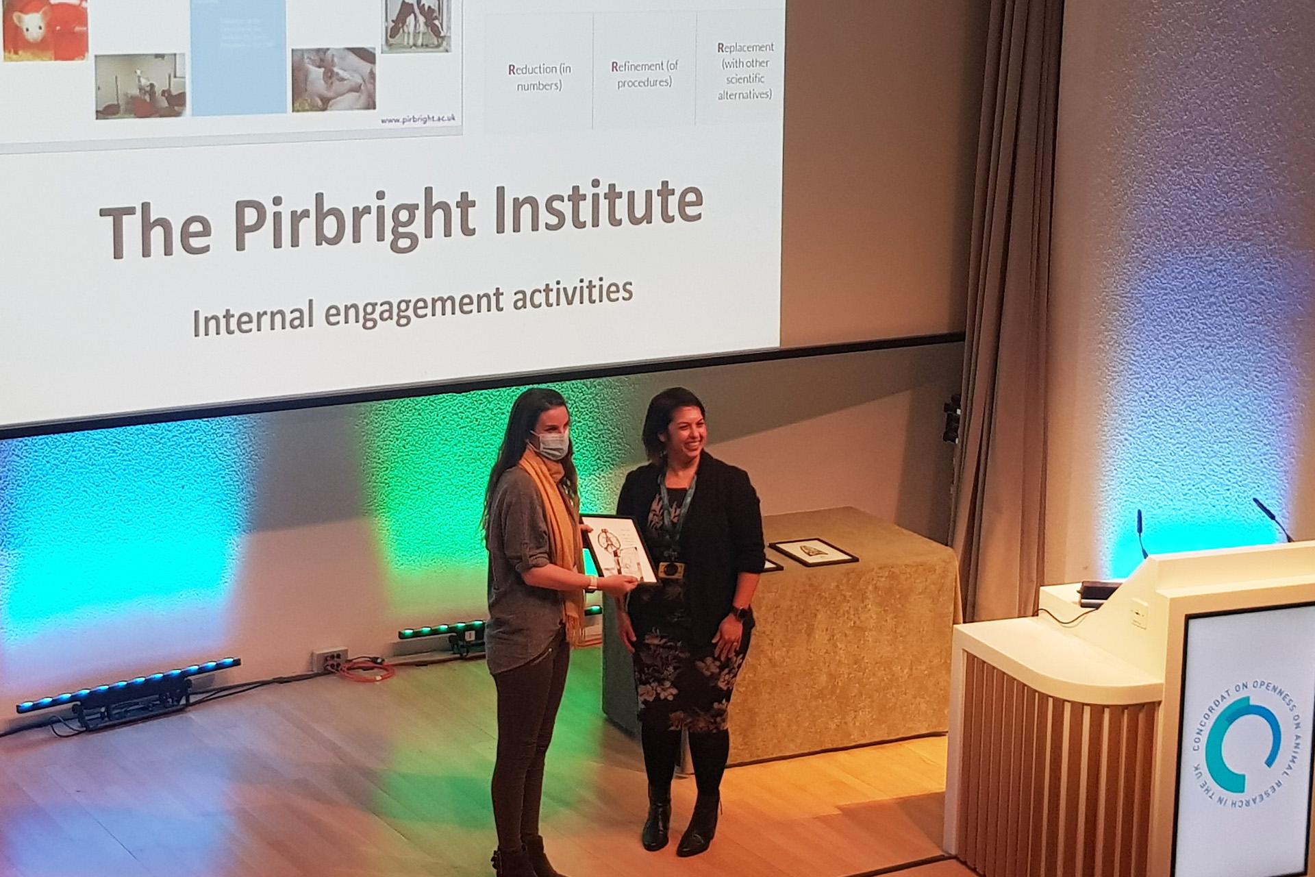 Two women on a lecture theatre stage (one with a face covering) holding a framed picture and glass trophy