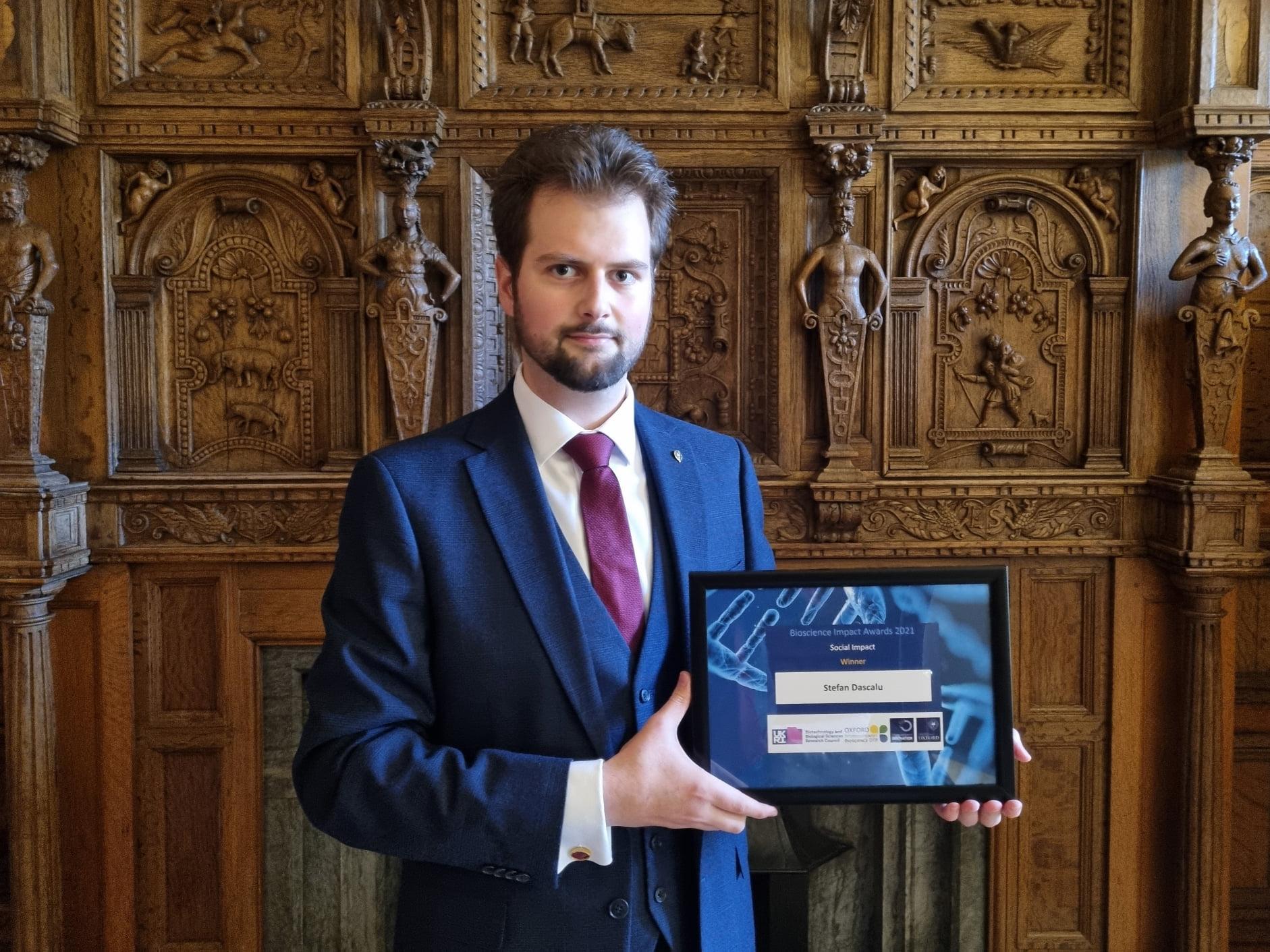 Stefan Dascalu in suit and tie, holds his Social Impact Award in front of wooden carved panel wall 