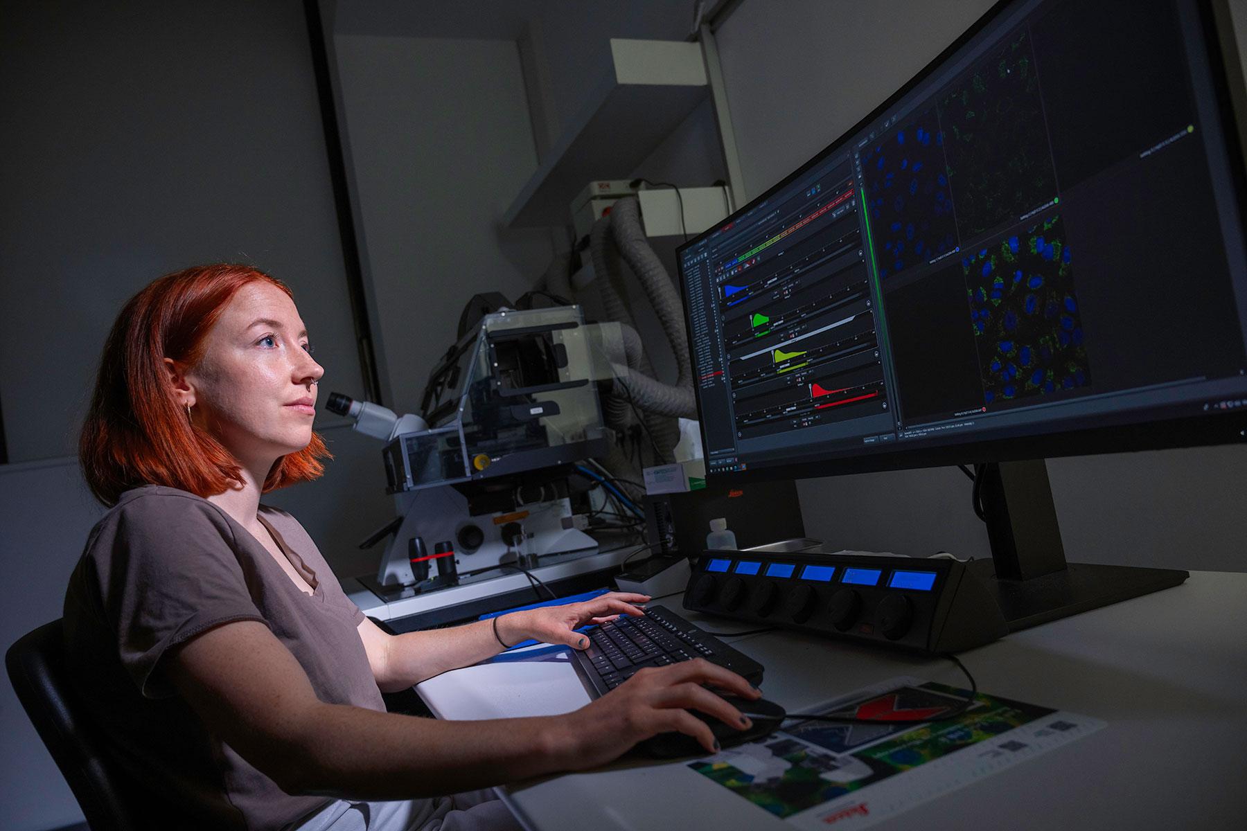 Scientist sat looking at two computer screens in a darkened room. On the screens are fluorescent images and graphs