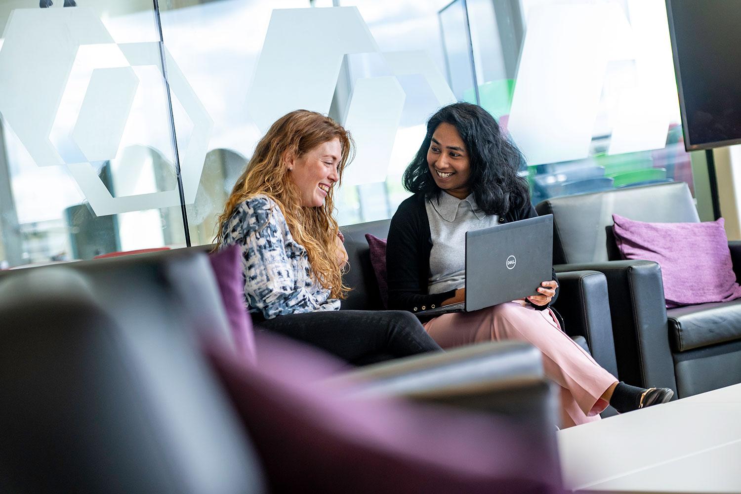 Two people sat on a sofa in front of clear glass that has hexagons on. They are both smiling. One has a laptop on their lap.