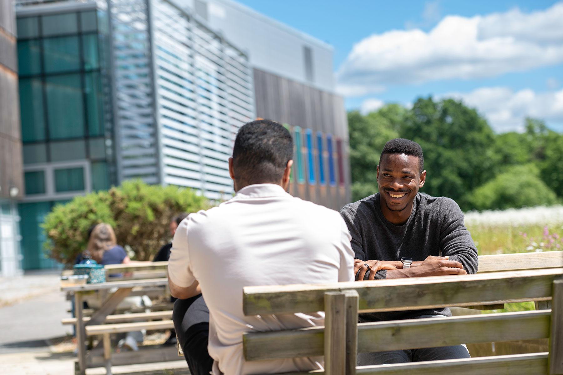 Two people outside sat at a picnic bench. One is facing the camera and smiling while looking at the other person who we only see the back of. It is a very sunny day with bright blue sky above and blurred out wildflowers in the background