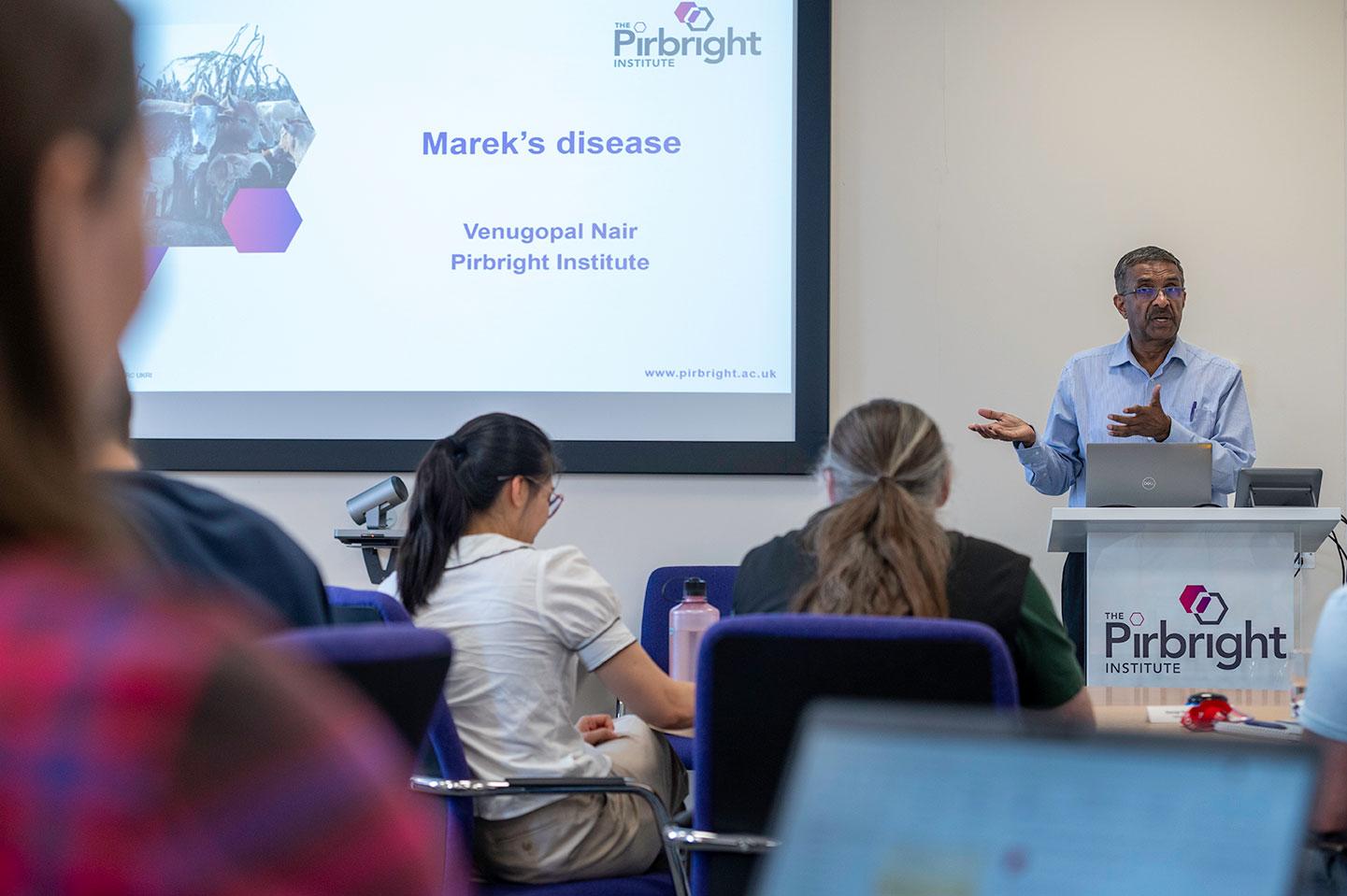 Person presenting at lecturn at the side of a large screen. On the screen a white slide with the pirbright institute logo contains black text that reads 'Marek's disease. Prof Venu Nair'