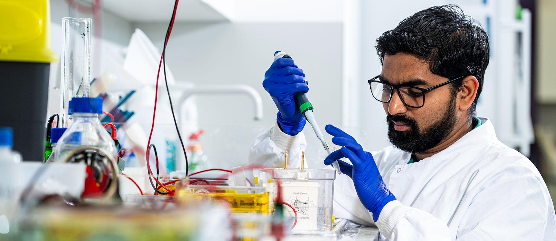 Researcher in white lab coat and purple gloved hands pipetting into an eppendorf tube 