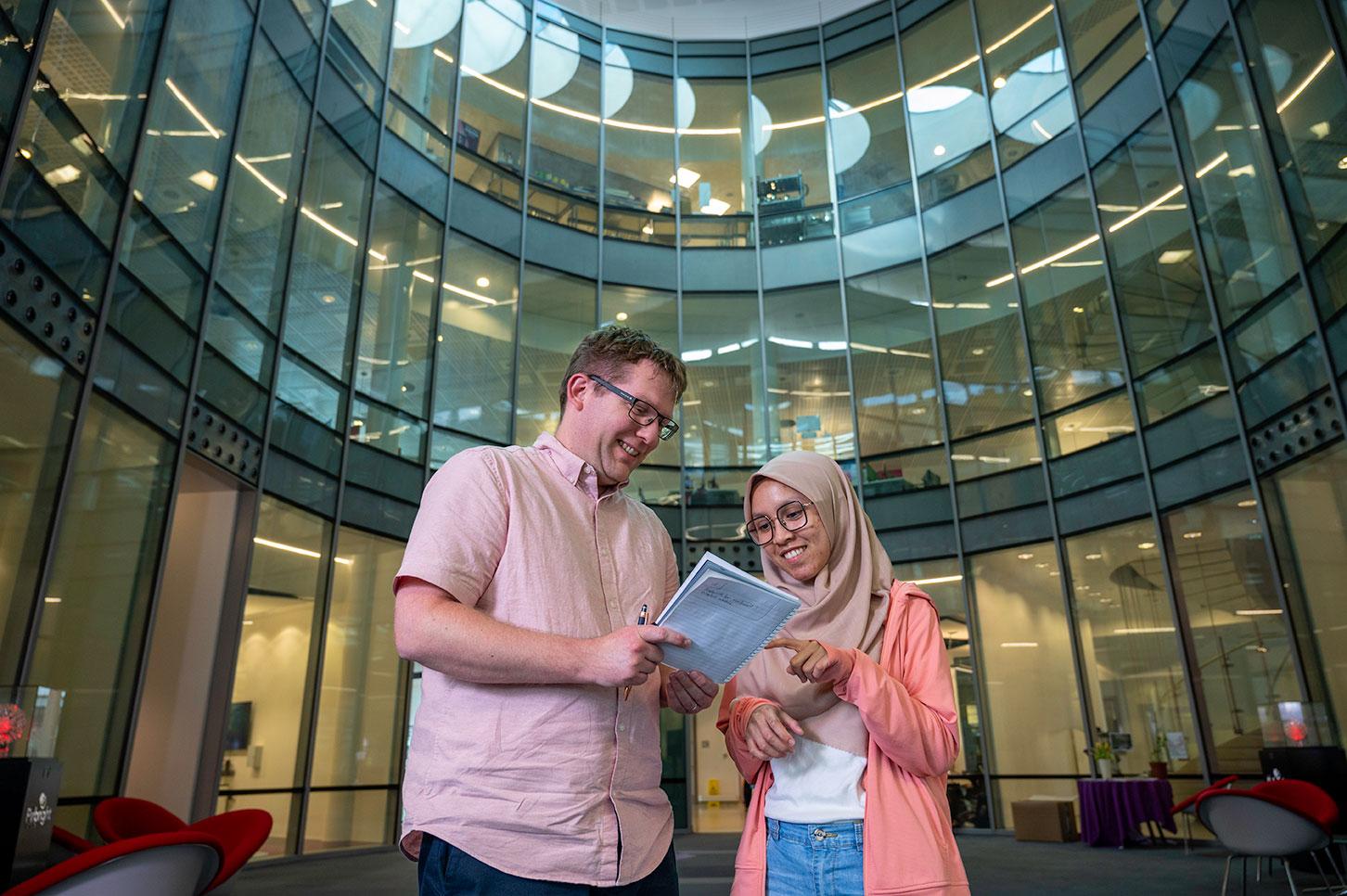 Two people stood in The Pirbriht Institute Plowright building atrium. They are looking at a notepad