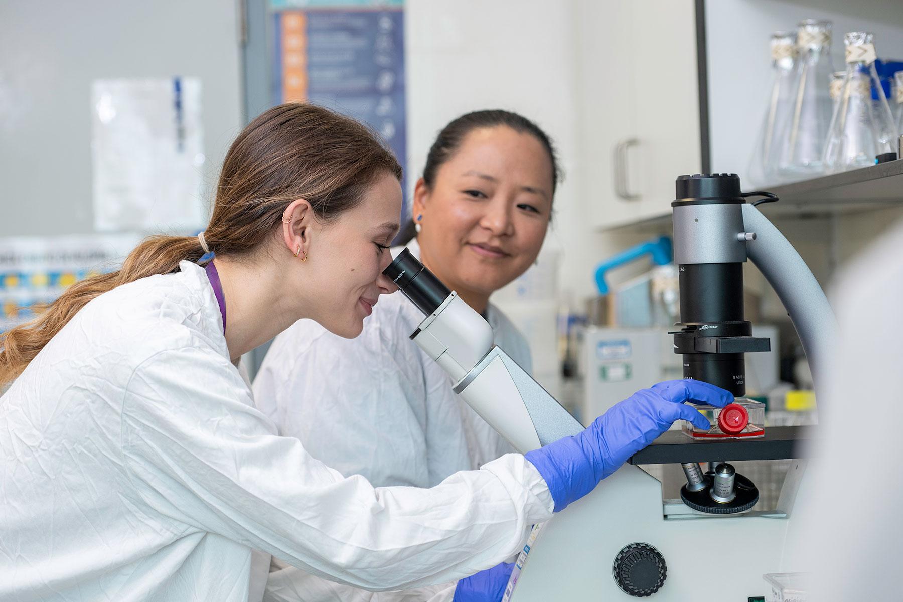 Two scientists in white lab coats. One is looking down a microscope with purple gloved hands hold a bottle under the microscope. The other person is in the background looking happily at the other