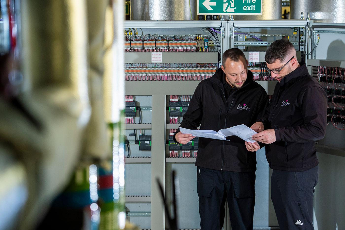 Two people stood looking a piece of paper in front a cabinet with electrical equipment