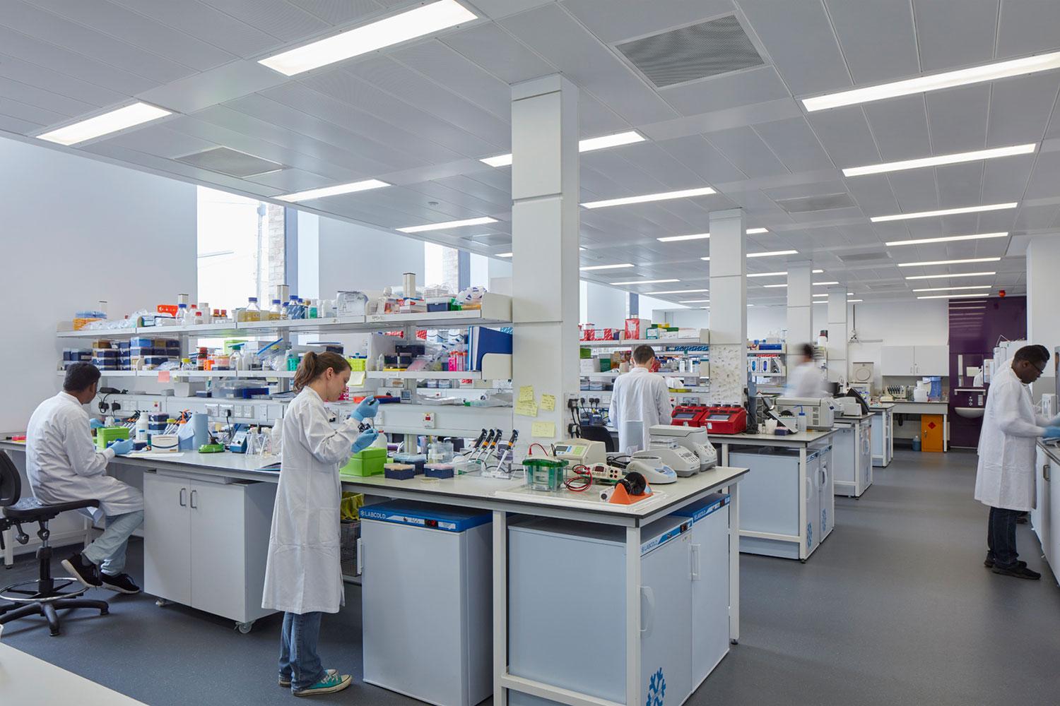 Large scientific lab with four rows of benches. It is full of equipment and boxes and people in lab coats moving about