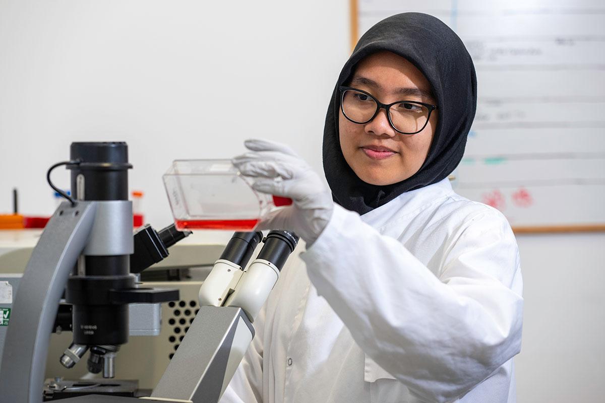 Scientist in white lab coat stood by microscope looking at bottle of red liquid