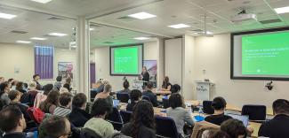 Delegates sat watching a presentation. There are two screens showing the same green title slide. A person stands at the lectern presenting