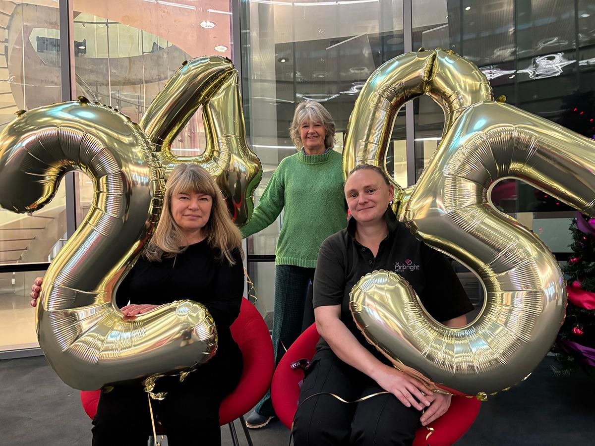 Linda, Alison and Louise. Linda is stood behind Alison and Louise with gold balloons 4 and 0 while Alison and Louise are sat. Alison on the left holds a gold 2 balloon while Louise holds a gold 5 balloon.