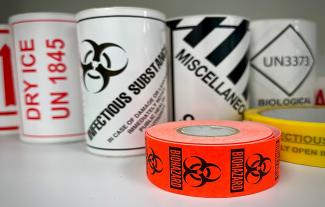 Seven rolls of tape on a shelf. One is white with infectious substance written on, one is red with biohazard written on. Other are white with "miscellaneous" and "biological" written on