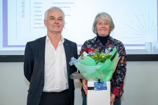 Prof Bryan Charleston and Dr Linda Dixon who is holding a box of flowers stood in front of a screen with agenda of symposium