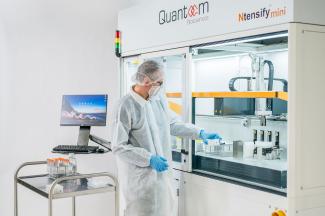 Researcher in a white lab coat, light blue gloves with a hair net and face mask stood in front of a trolley with plastic tubes on. The researcher is placing a small plastic box into a cabinet that is branded with Quantoom Biosciences Ntensify 