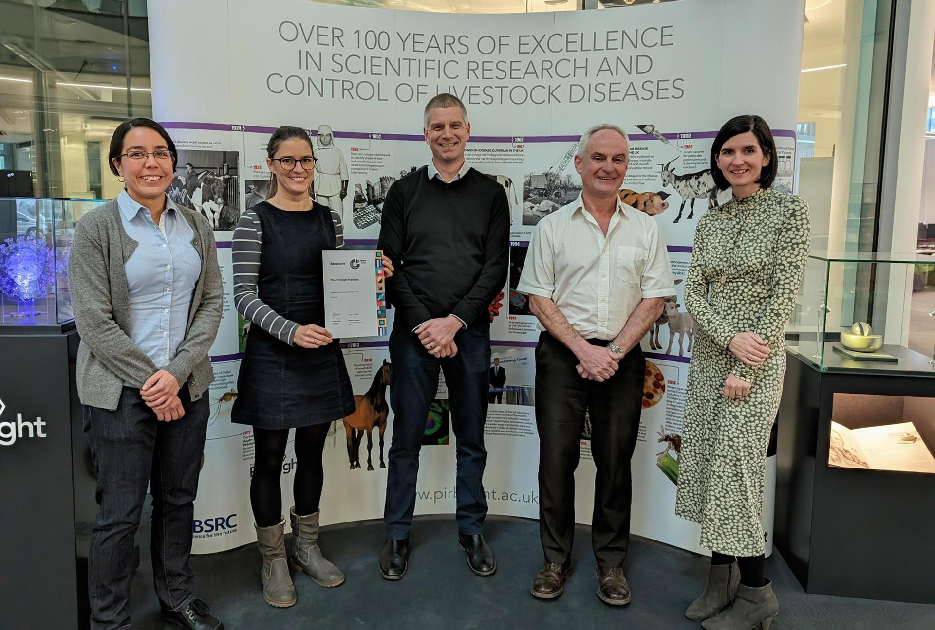 Group of five people from Pirbright in a line smiling in front of a large white poster. One is holding a certificate.