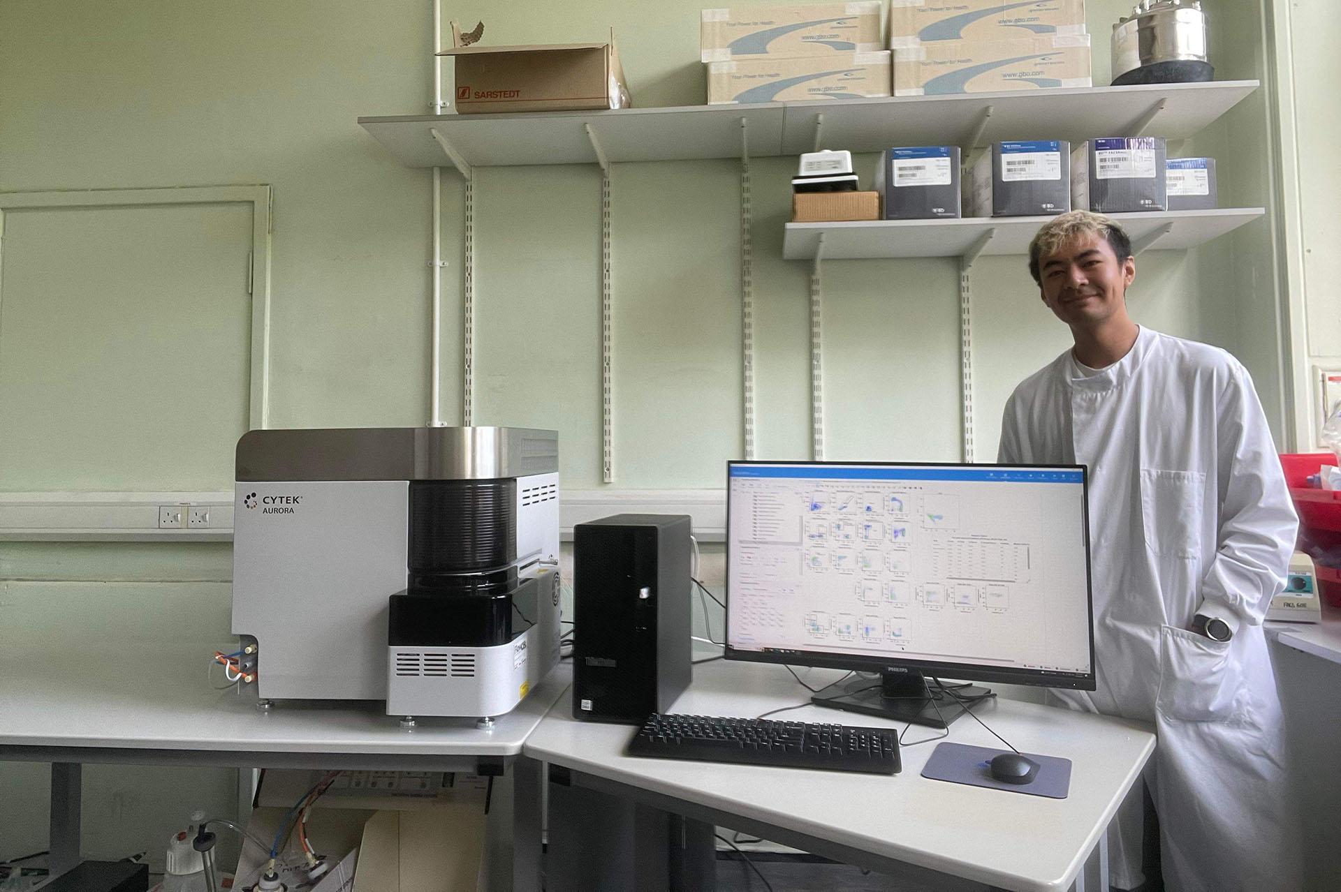 Young scientist Chis stood in a lab wearing a lab coat. Graphs are shown on the computer screen.