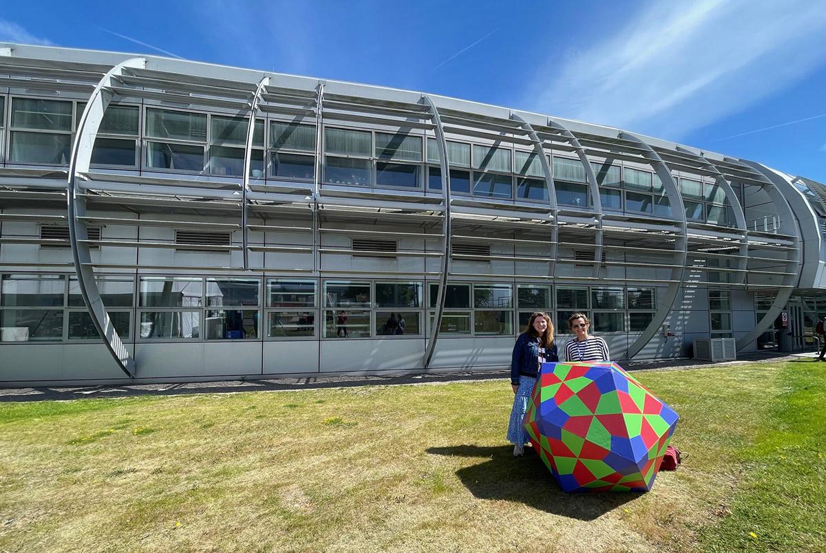 Two people with a giant red, green and blue FMDV model stood in front of a building