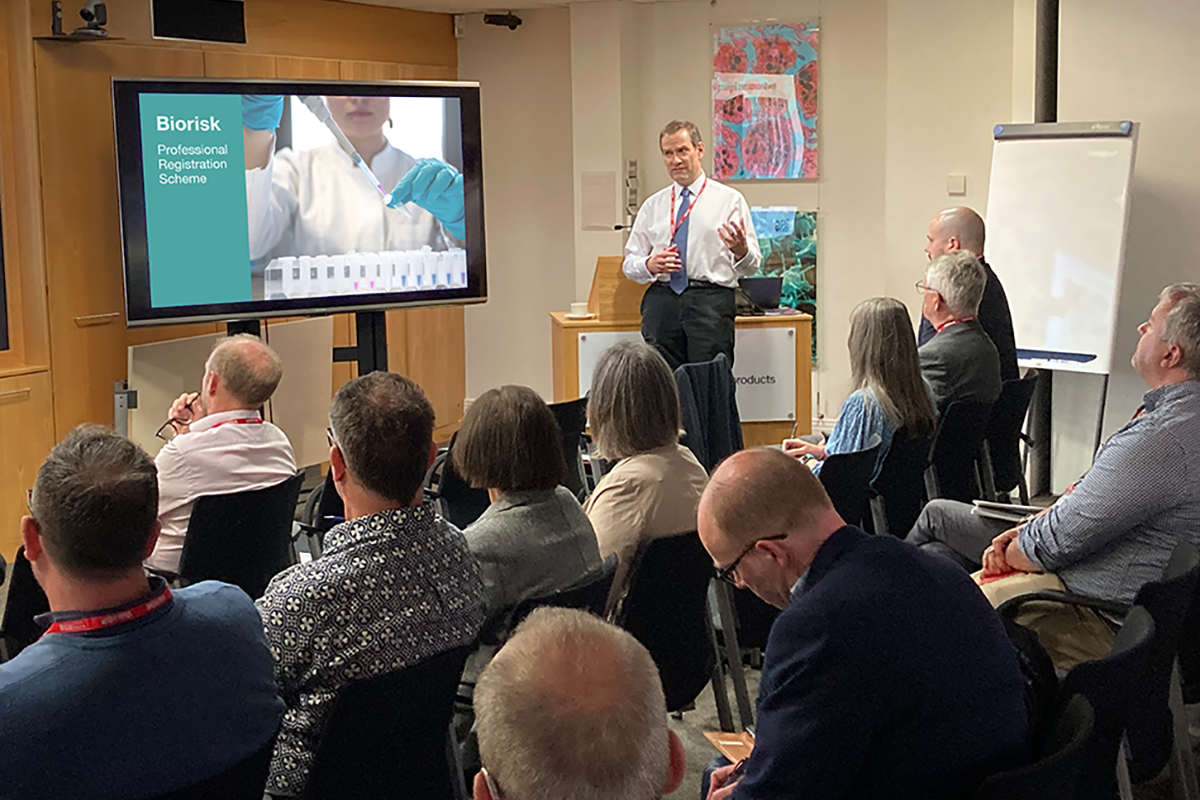 Person stood presenting in front of seated group. On the tv screen is a slide of someone pipetting with the title biorisk