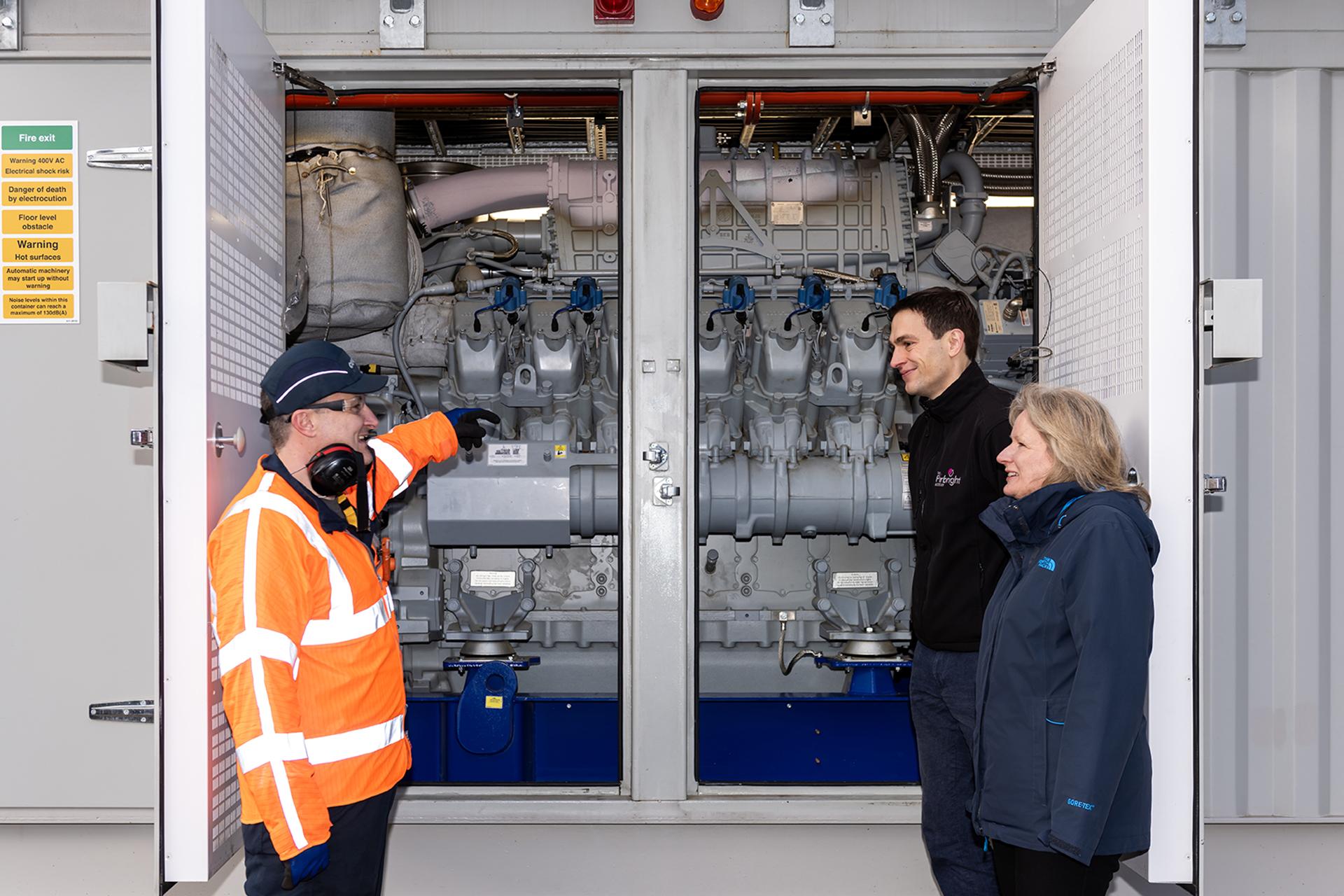 A person in a fluorescent orange jacket pointing at electrical parts on the left with two people on the right in jackets smiling